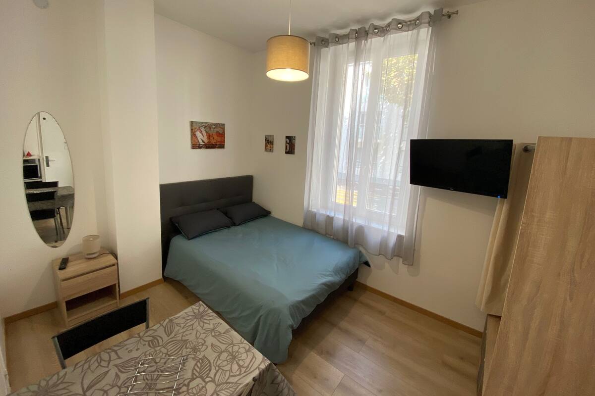 A bedroom with a bed, a bedside table, a mirror, a TV, and a window with curtains.