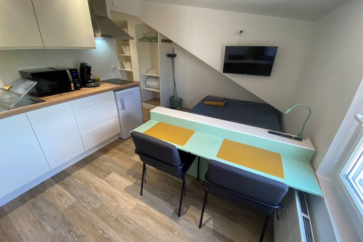 A compact kitchen with a microwave, coffee maker, and a small refrigerator. There is a dining area with two chairs and a table, and a TV mounted on the wall.