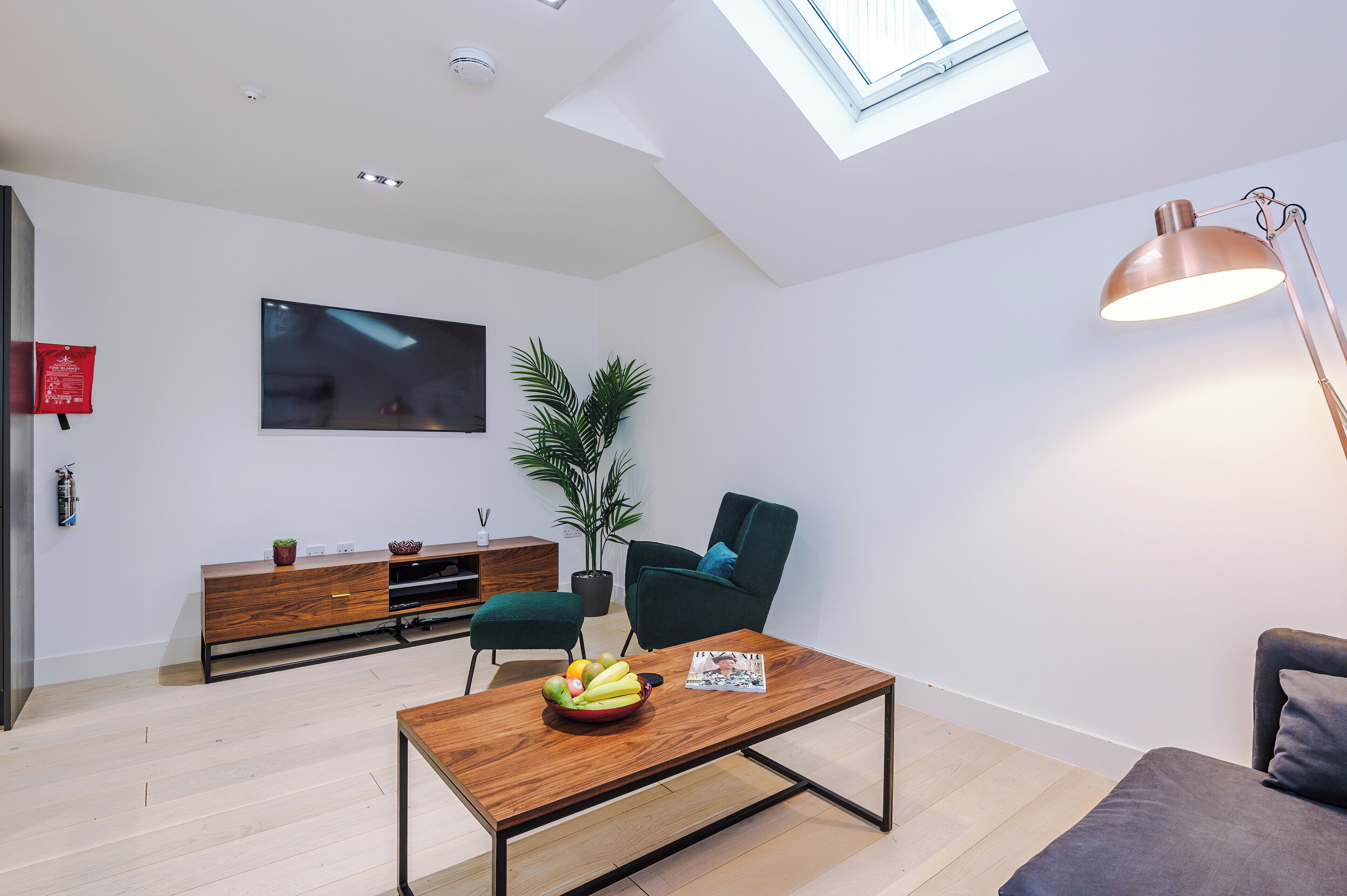 A modern living room with a wooden coffee table, a green armchair, a sofa, a TV on the wall, and a skylight.