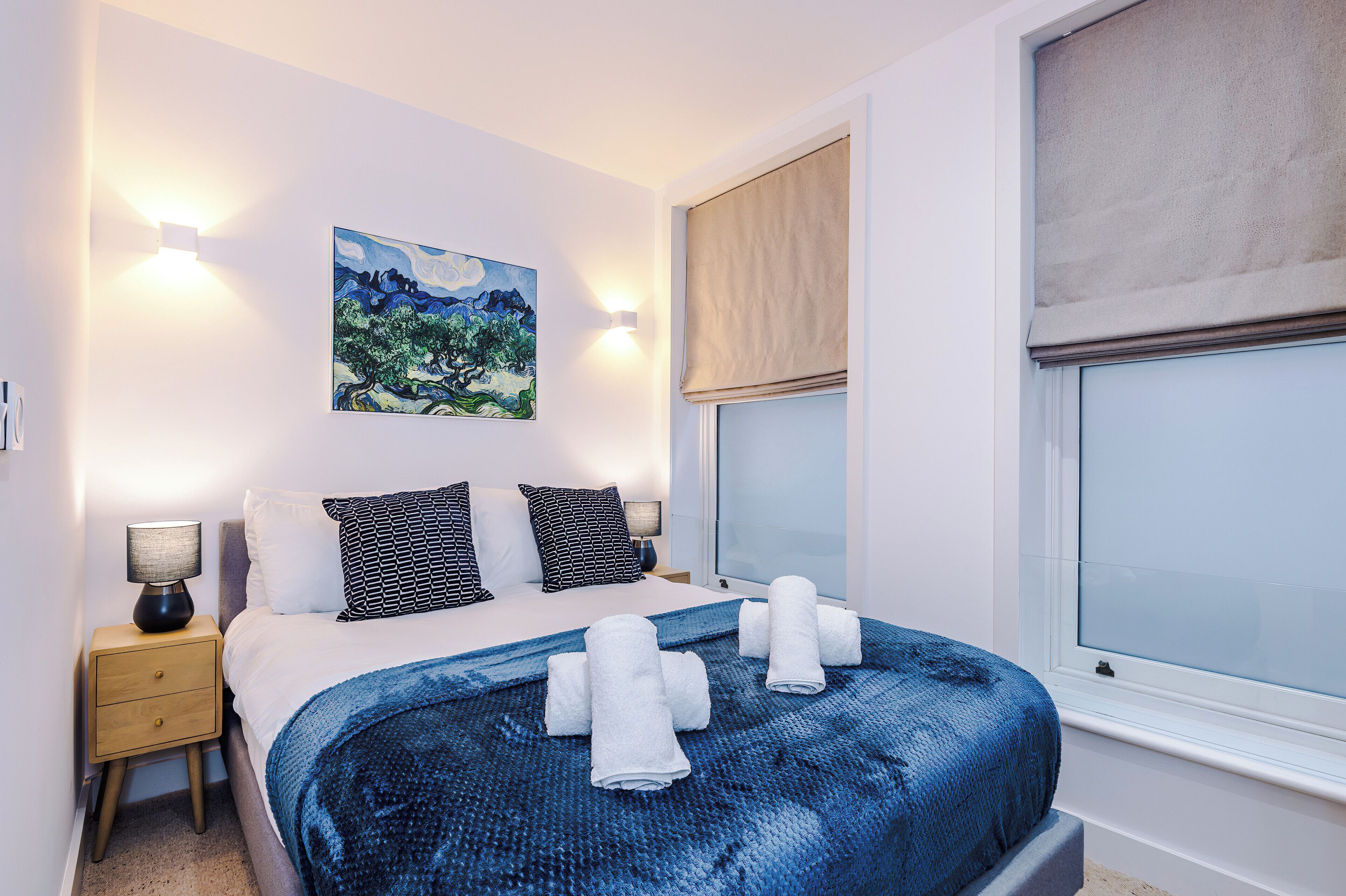 Cama bem arrumada com manta azul texturizada, duas luminárias de apoio, um quadro na parede e uma janela com persiana bege.
