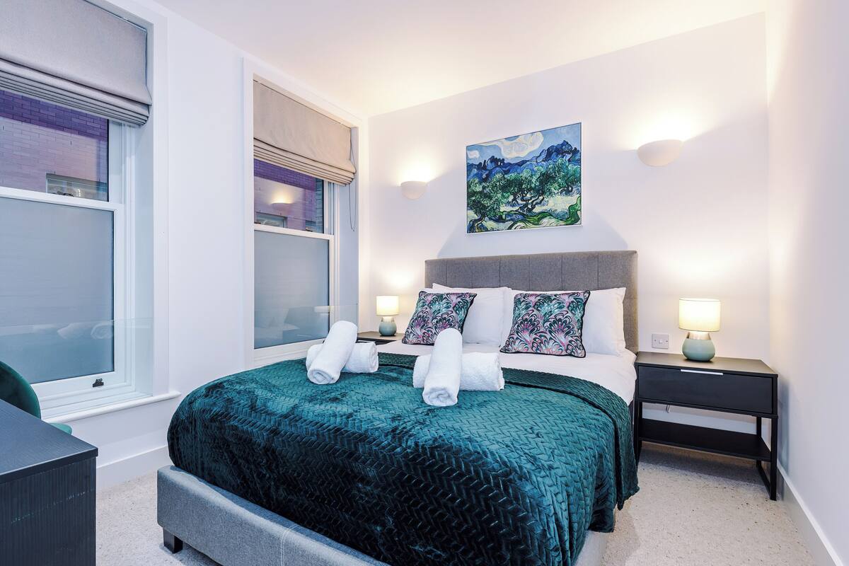 Ein modernes Schlafzimmer mit einem großen Bett, zwei Nachttischen, einem Beistelltisch und einem Fenster mit Jalousien.