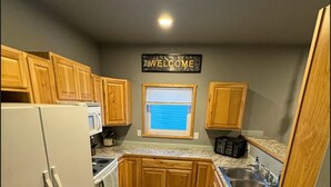 A compact kitchen with wooden cabinets, a white refrigerator, a microwave, and a sink. A welcome sign is visible on the wall. - Modern 2-bed with full kitchen, in-unit laundry, and off-street parking. Near downtown Fairbanks. (Fairbanks)