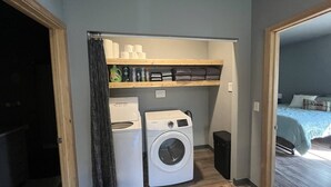 A compact laundry room with a washing machine and dryer, shelves with laundry supplies, and a bed in the adjacent room. - Modern 2-bed with full kitchen, in-unit laundry, and off-street parking. Near downtown Fairbanks. (Fairbanks)