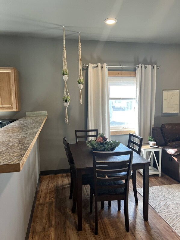 A dining area with a wooden table and chairs, a kitchen with a wooden countertop, and a window with curtains.