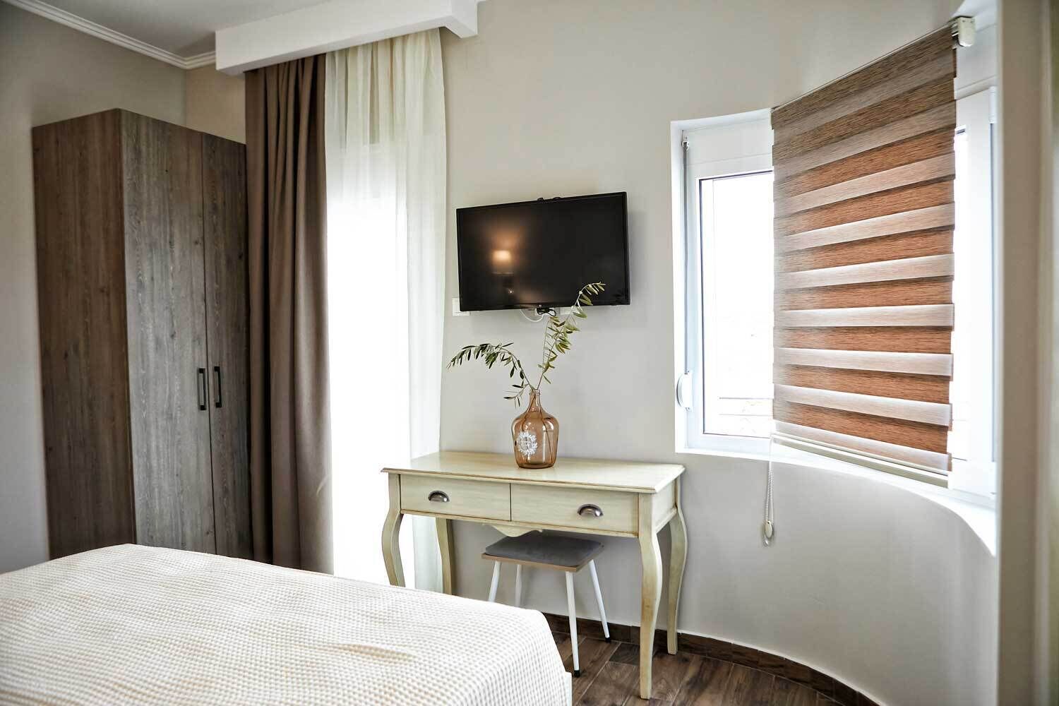 Quarto com cama, escrivaninha com um vaso, televisão e janela com persianas.