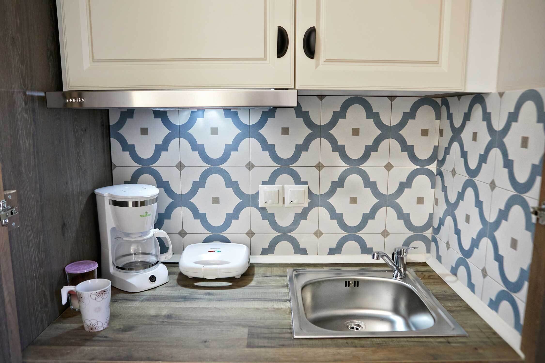 Una cocina compacta con gabinetes blancos, un lavamanos de acero inoxidable, una cafetera, una balanza y un salpicadero con diseño.