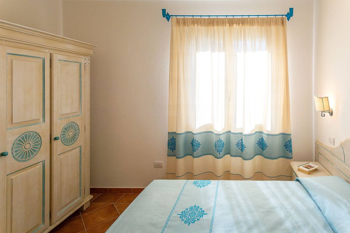 Un dormitorio con cama, mesita de noche, armario y ventana con cortinas.