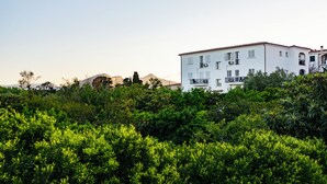 Een wit gebouw met meerdere balkons, omgeven door weelderige groene beplanting en bergen op de achtergrond.