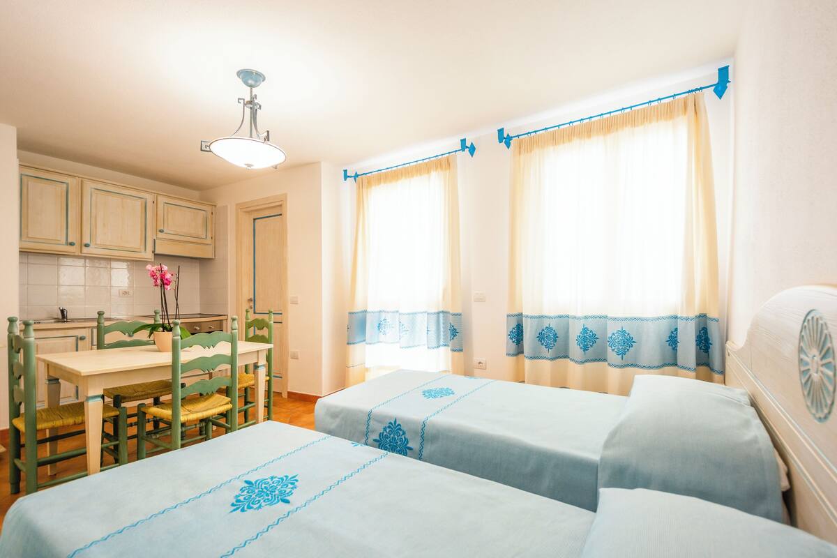 Una habitación con dos camas, una mesa de comedor con sillas, una zona de cocina y una ventana con cortinas.