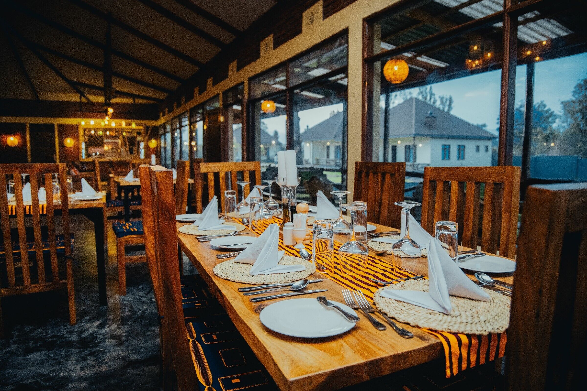 A restaurant with wooden tables set for dining, large windows, and a view of a house outside.