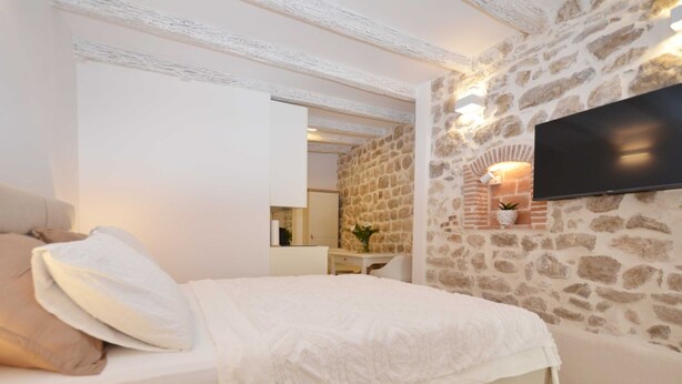 Een slaapkamer met een stenen muur, een flatscreen televisie boven een open haard, een bed met een witte dekbedovertrek en een nachtkastje met een lamp.