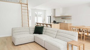 Ein modernes Wohnzimmer mit einem Ecksofa, einer Holzleiter, einem Essbereich mit Holztisch und -stühlen sowie einer Küche im Hintergrund.