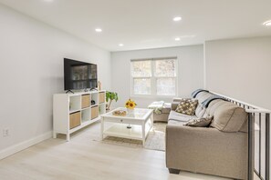 Un salon moderne avec un canapé blanc, une table basse, un téléviseur posé sur un meuble blanc et une fenêtre donnant sur des arbres.
