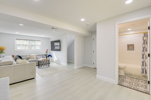 A spacious living room with a sofa, coffee table, and a large window. There is a door leading to a bathroom with a toilet and bathtub. - Near Top Attractions: Deck Dining at Severn Home! (Severn)