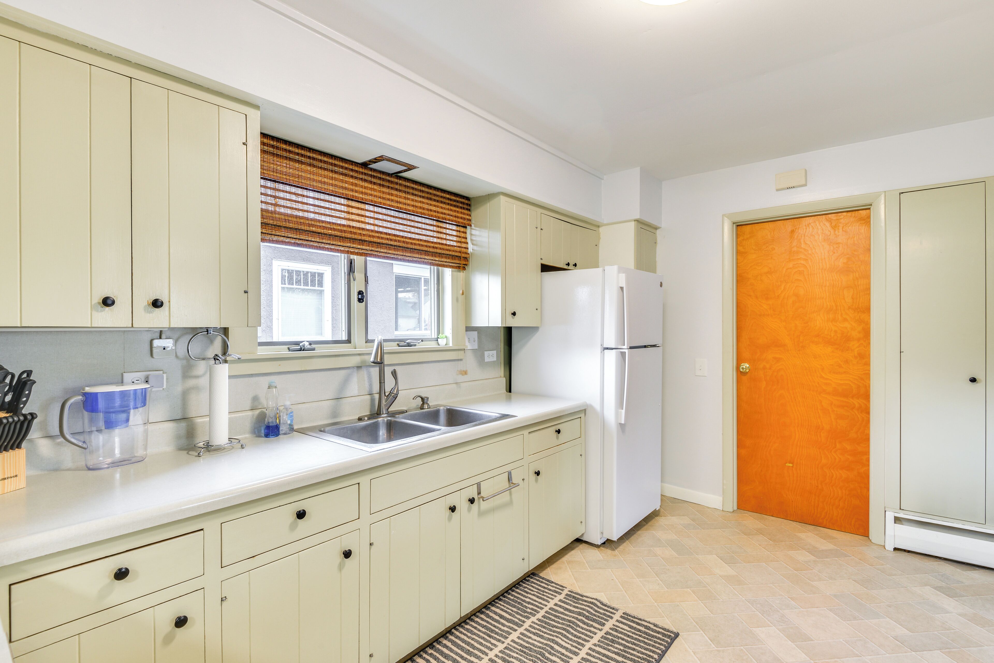 A kitchen with light-colored cabinets, a white refrigerator, a sink, and a window with blinds.