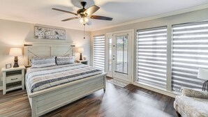 Ein Schlafzimmer mit Bett, Nachttischen, Deckenventilator und großen Fenstern mit Jalousien.