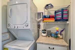A laundry area with a front-loading washing machine, a shelf with folded towels, a laundry basket, and cleaning supplies. - West Bay View: Penthouse Condo in Traverse City (Traverse City)