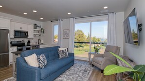 Un salon moderne avec un canapé bleu, un fauteuil beige et une grande fenêtre donnant sur un jardin et des arbres.