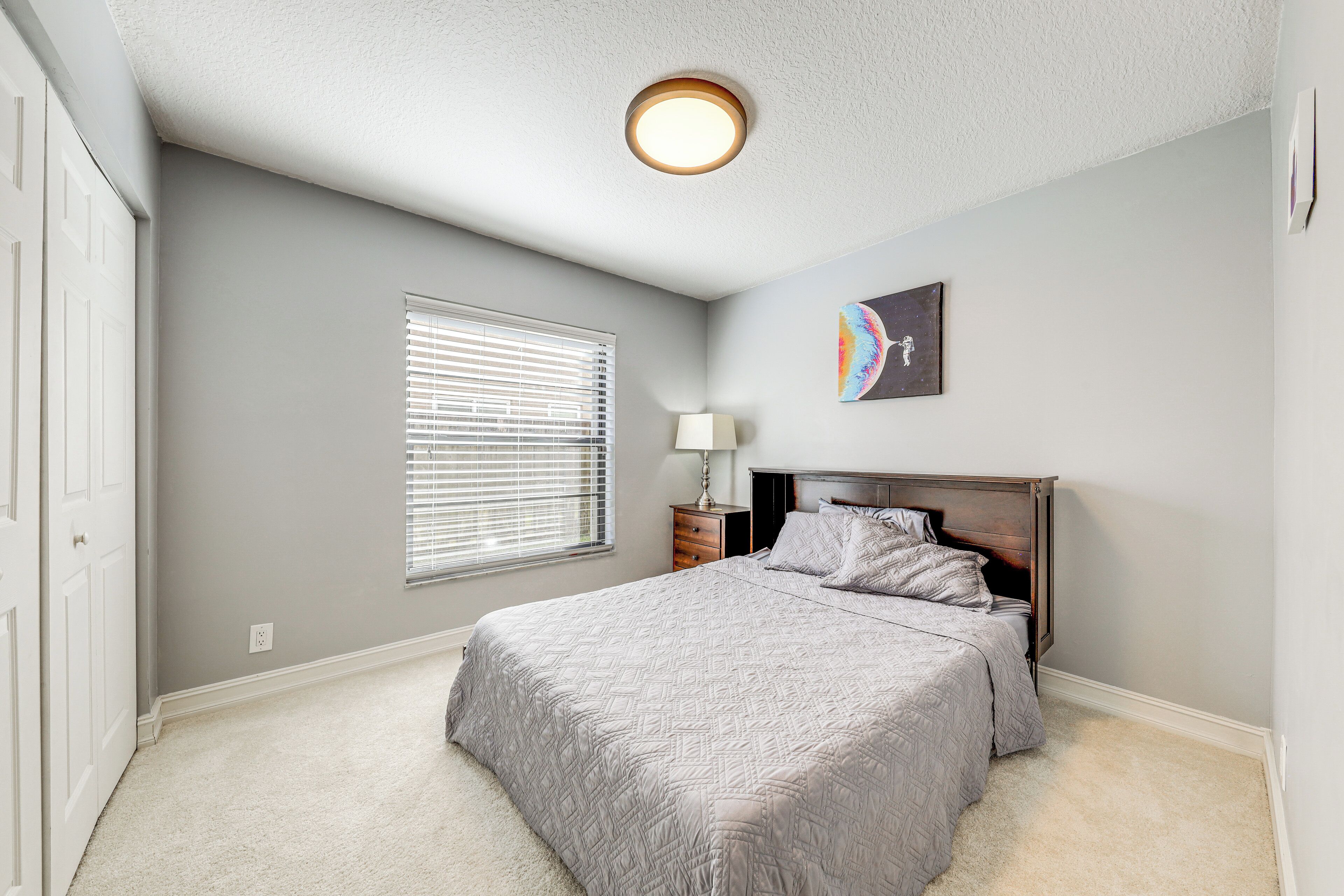 A bedroom with a bed, a nightstand, a lamp, a window with blinds, and a ceiling light fixture.