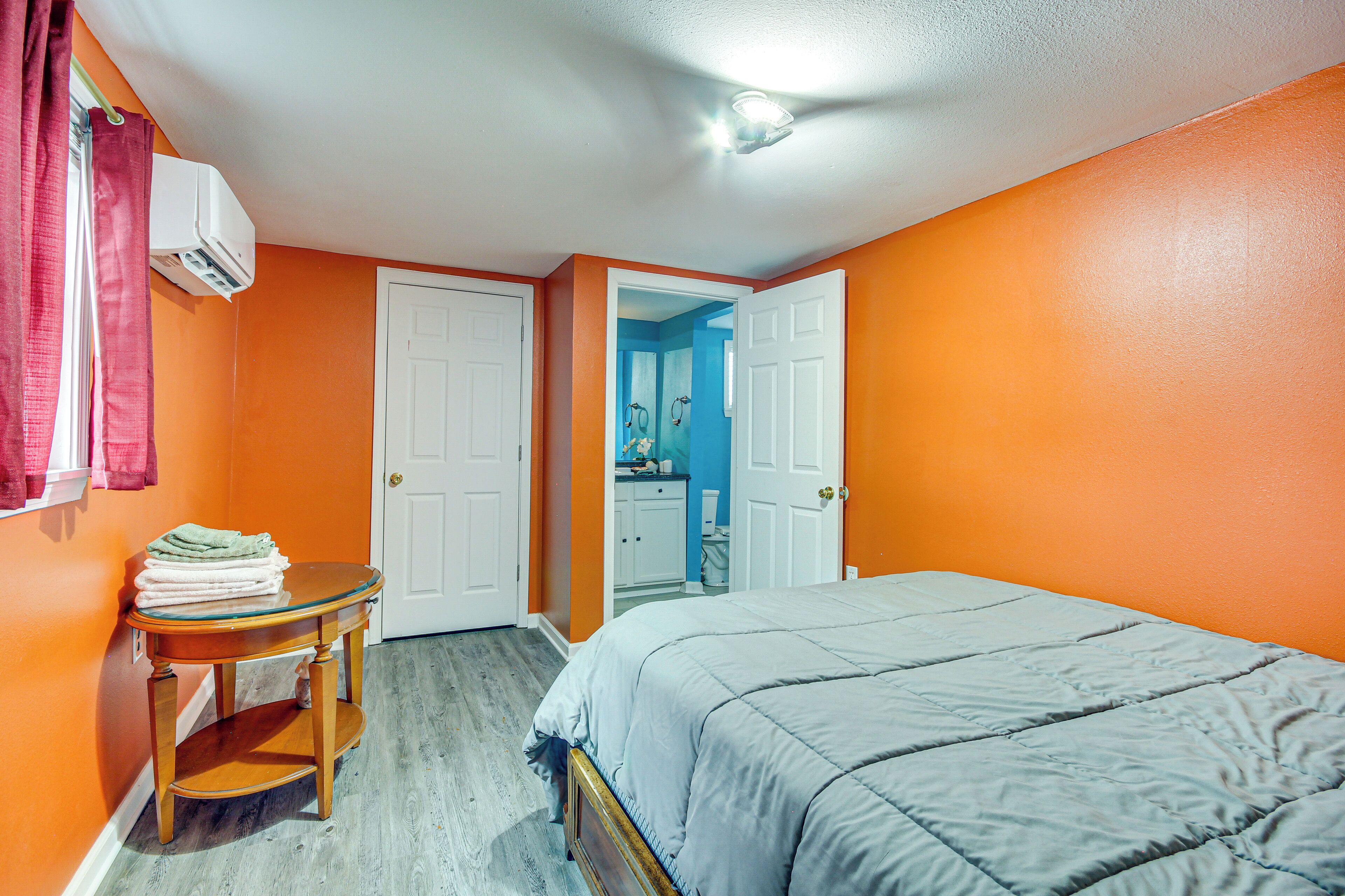 Quarto com parede laranja, cama com colcha cinza, mesa de cabeceira de madeira com uma pilha de toalhas dobradas e porta branca que dá para um banheiro.