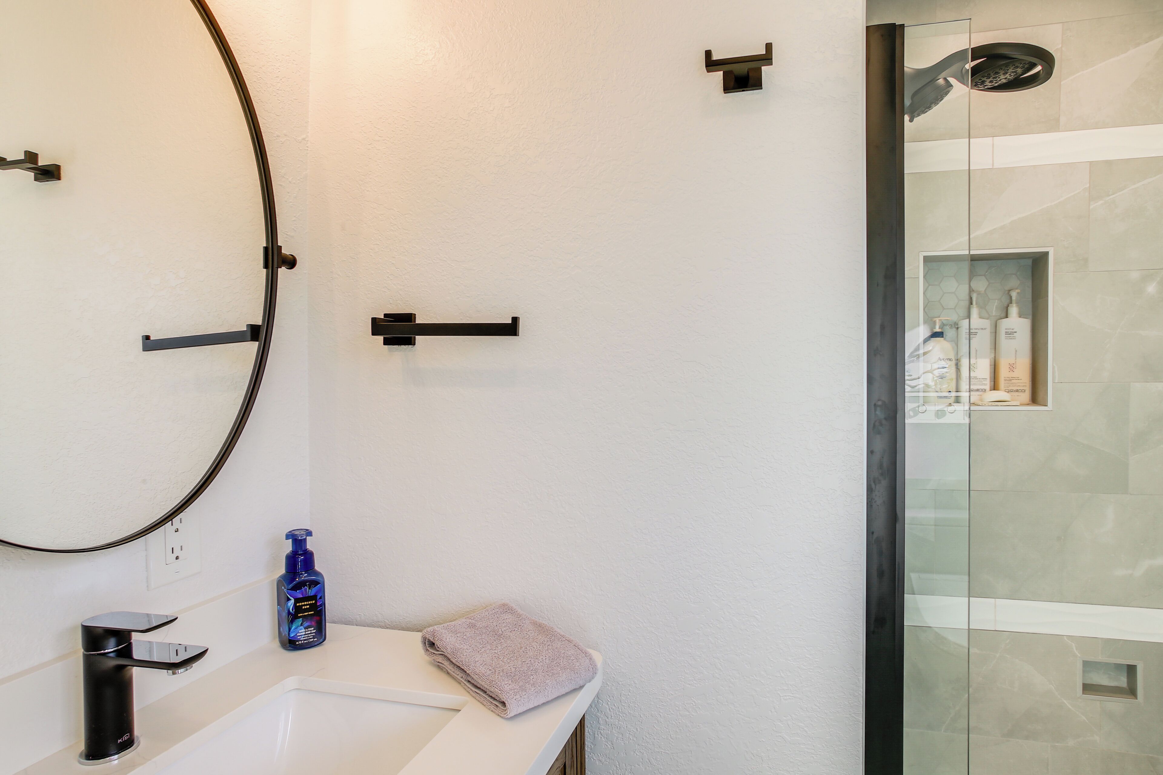 A modern bathroom with a white sink, a round mirror, a handheld showerhead, and a glass-enclosed shower.