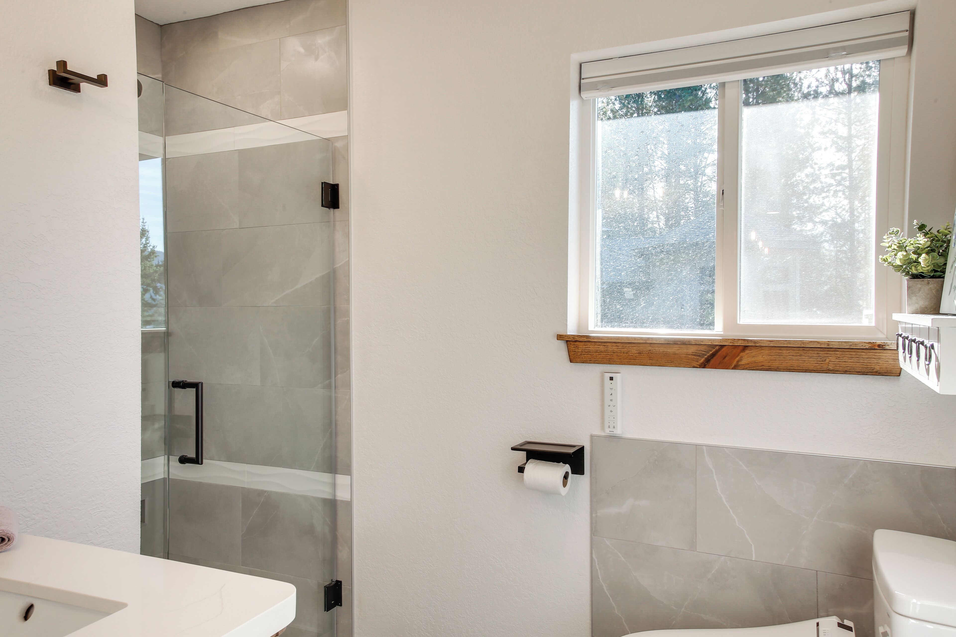 A modern bathroom with a glass shower enclosure, a toilet, and a window with a view of trees.