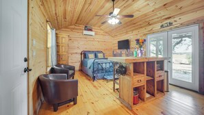 Ein Interieur einer Holzhütte mit Holzbalkendecke, einem Bett mit blauer Bettwäsche, einem Essbereich mit Tisch und Stühlen sowie einem großen Fenster.
