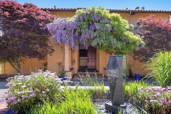 A yellow building with a purple flowering vine, a stone pillar, and various plants.