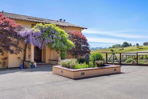 A building with a tiled roof, a garden with various plants, and a fenced area with a view of the countryside.