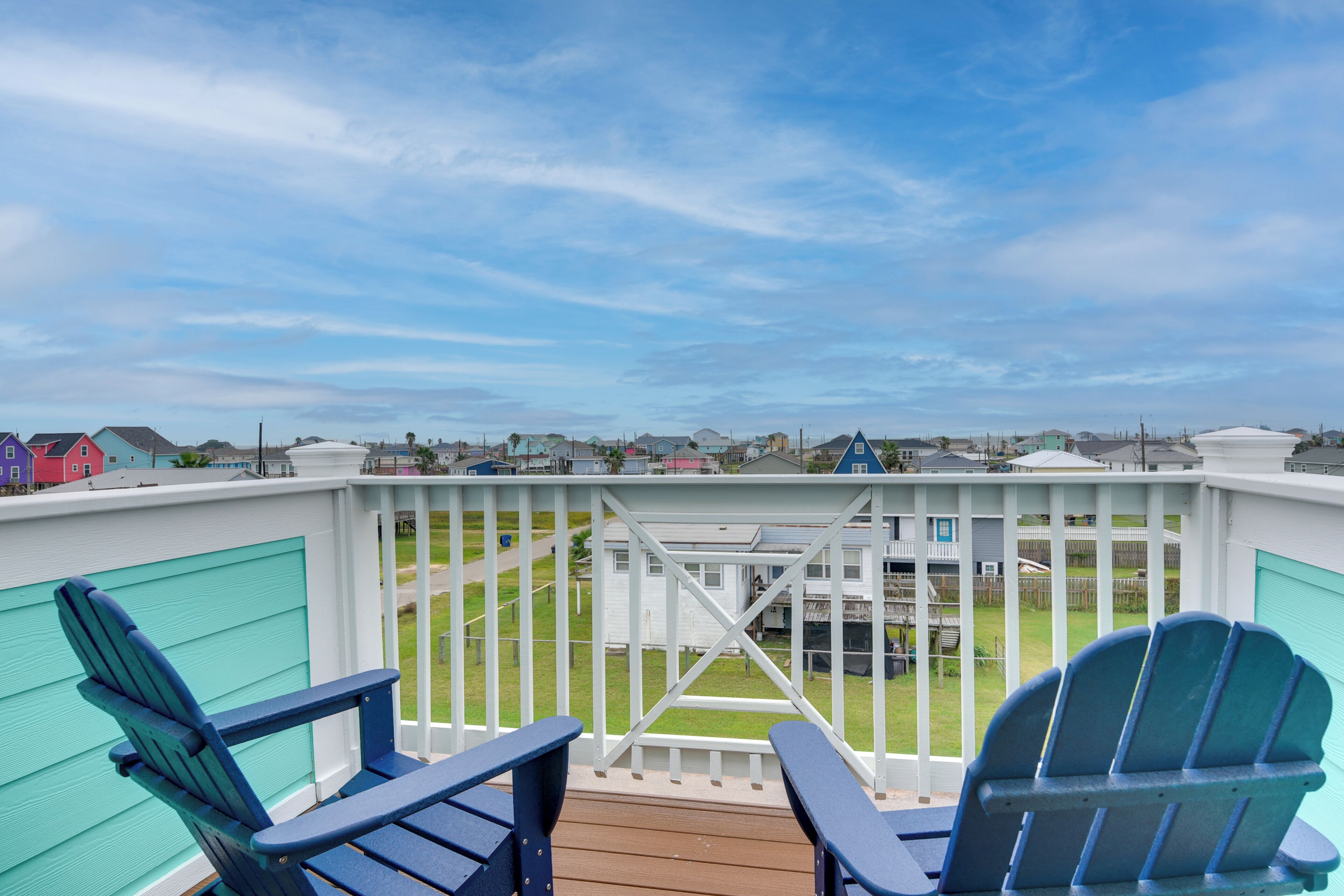 A deck with two blue Adirondack chairs, a white railing, and a view of a residential area with houses and a grassy field.