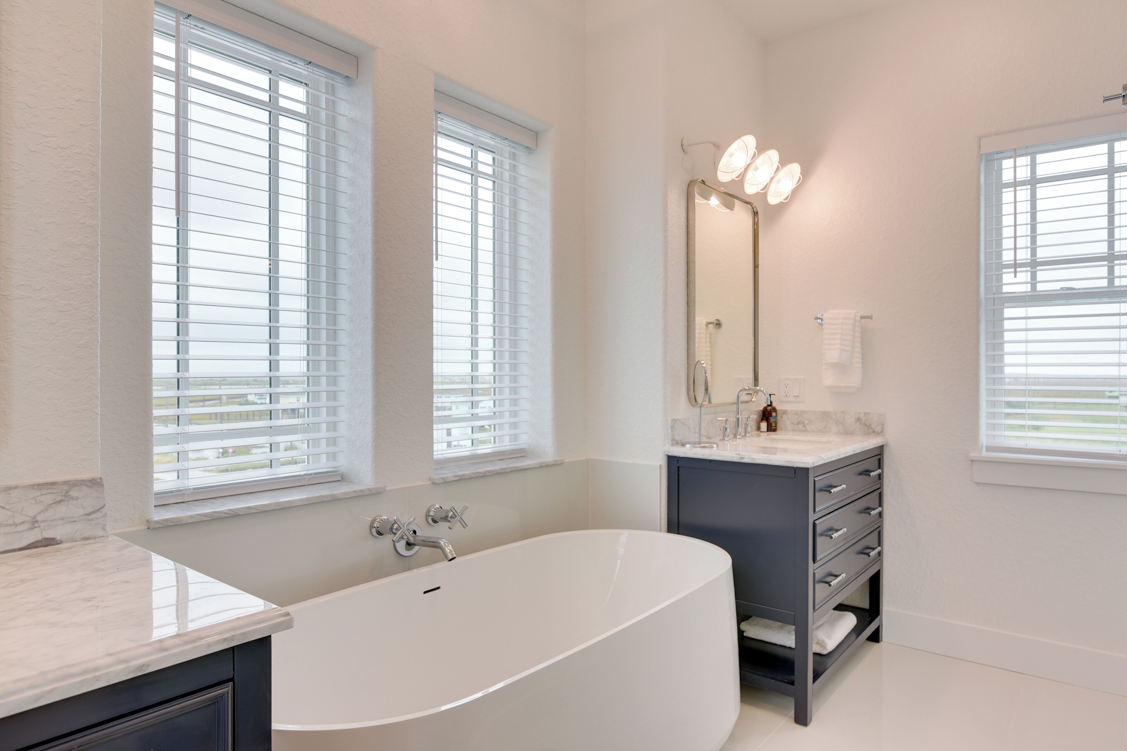 A modern bathroom with a freestanding bathtub, a vanity with a mirror, and two windows with blinds.