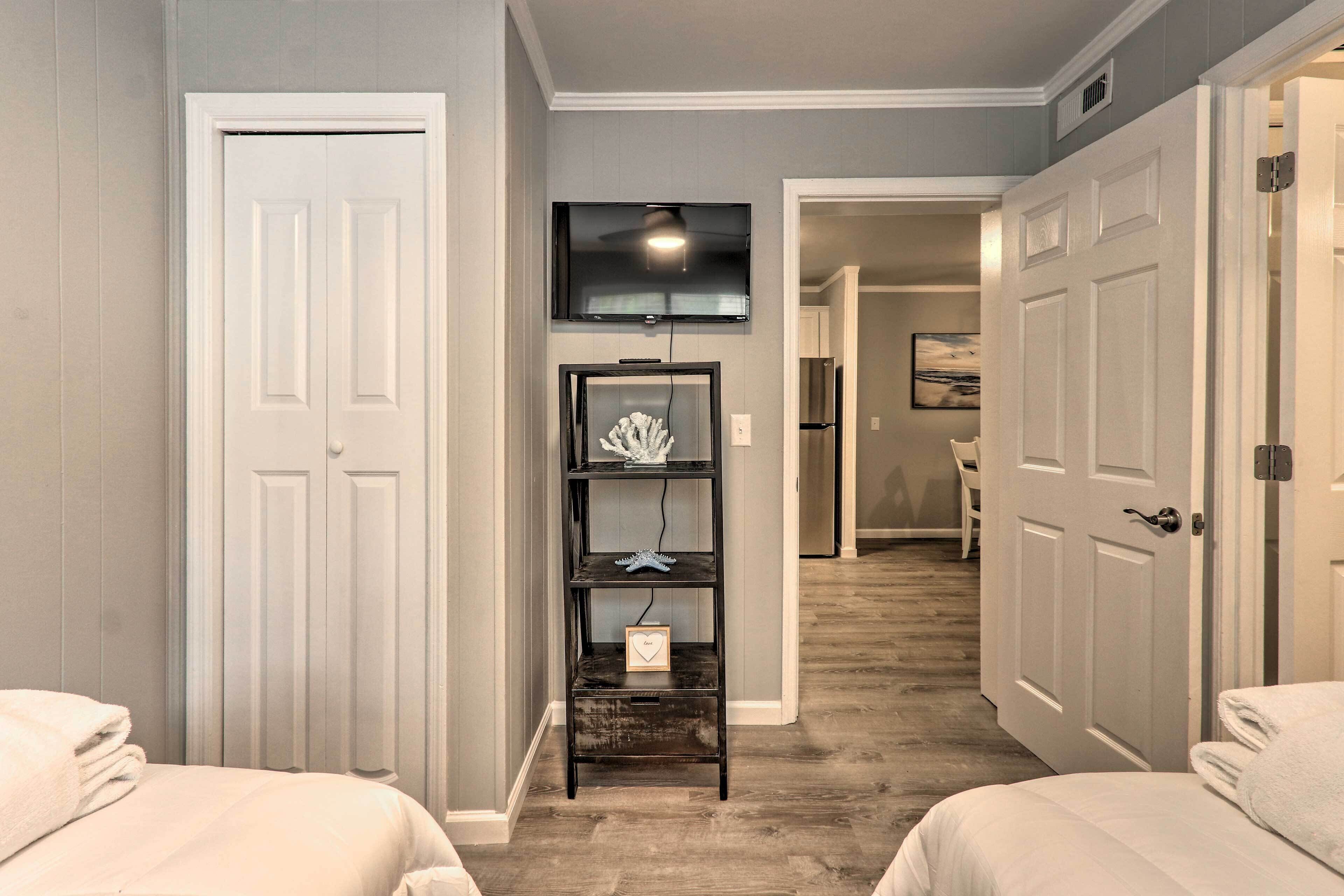 A hotel room with two beds, a TV mounted above a wooden shelf, and a hallway leading to a kitchen.