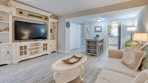 A living room with a large flat-screen TV, a beige sofa, a coffee table, and a kitchen area visible through an open door. - Walk to Ocean - Townhome w/ Patio in Myrtle Beach! (Myrtle Beach)
