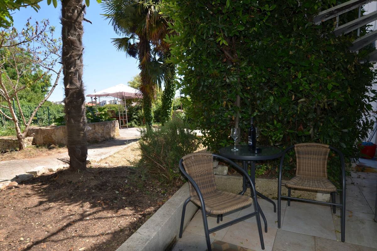 An outdoor patio area with two wicker chairs and a table, surrounded by lush greenery and a palm tree.