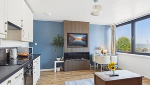A modern kitchen with a built-in oven, a dining area with a table and chairs, a fireplace, and a large window with a view. - Harbour Hideaway - 1 Bedroom Apartment - Saundersfoot (Saundersfoot)