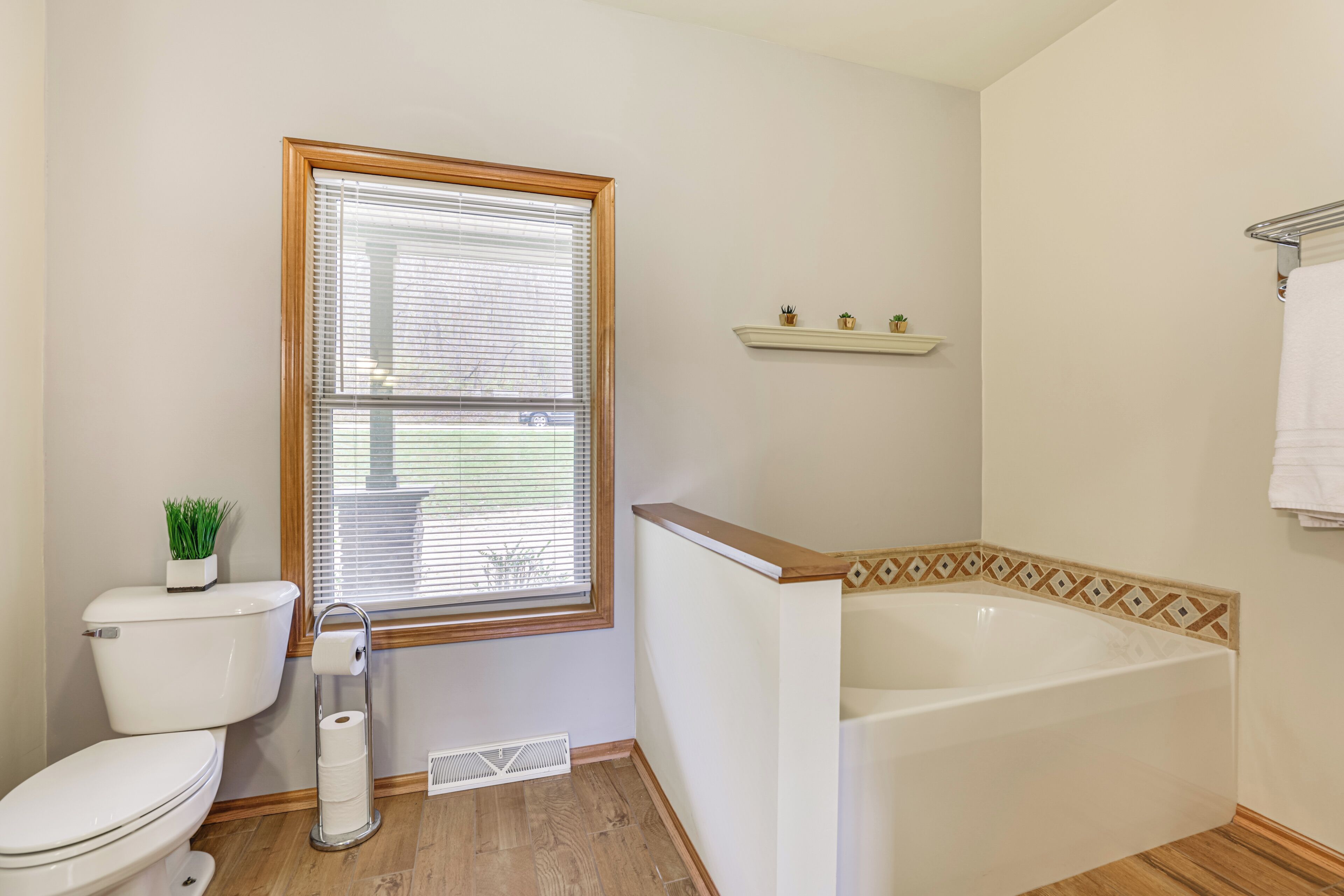 Ein Badezimmer mit Toilette, einem Fenster mit Jalousien, einer Badewanne und einem Pflanzenregal.