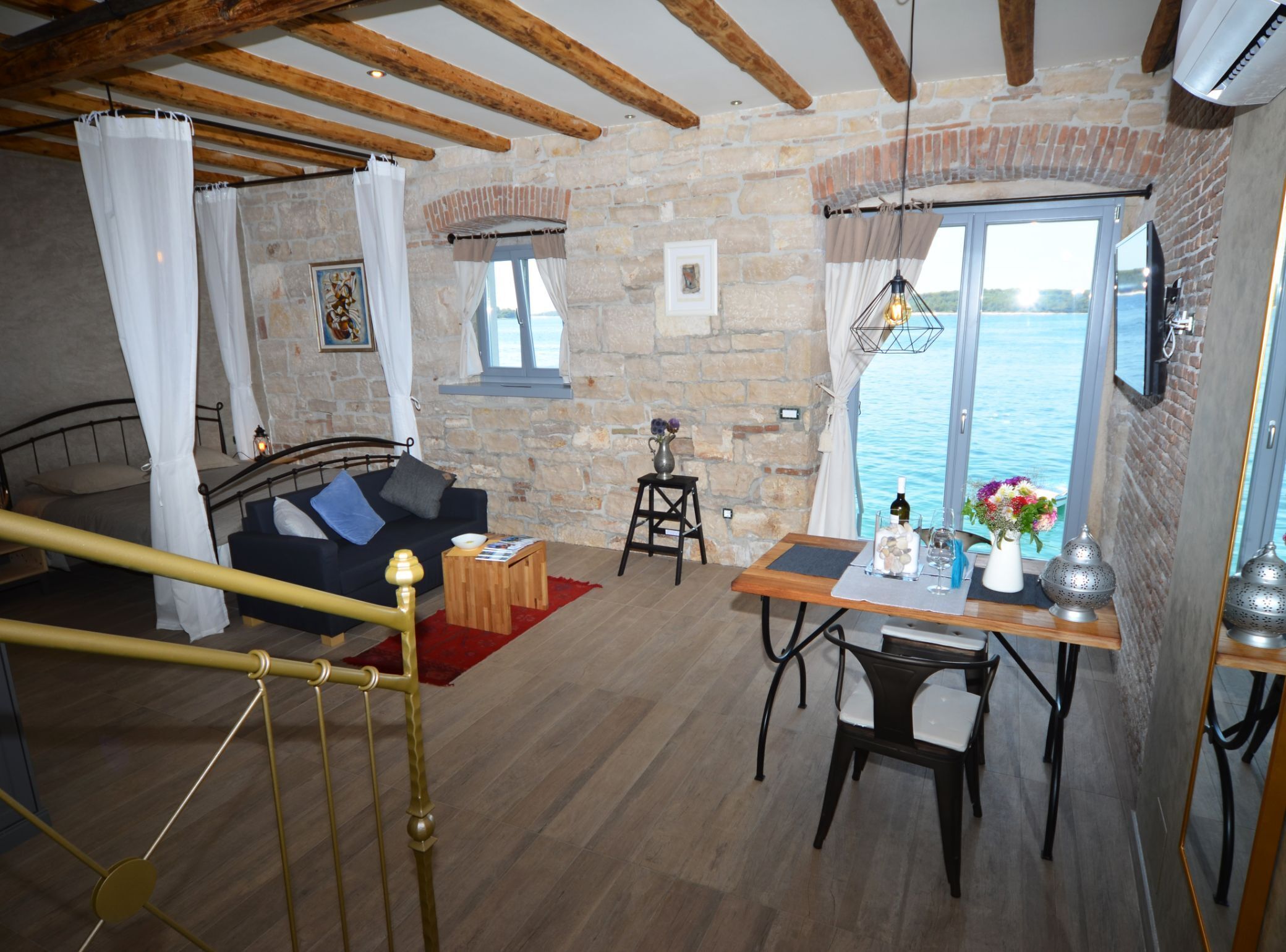 Una habitación acogedora con cama, sofá, mesa de comedor con sillas, una mesita con un jarrón y copas de vino, y vista al mar a través de amplias ventanas.