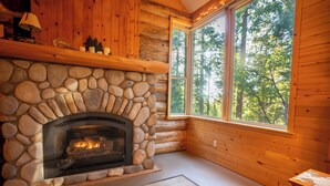 A stone fireplace with a log fire, wooden walls, a large window with a forest view, and a wooden table with a lamp.