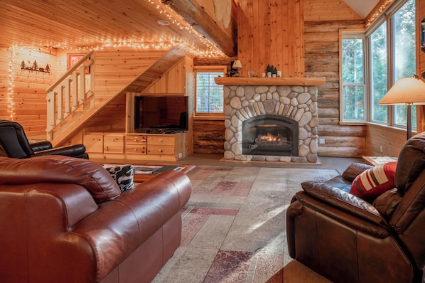 A log cabin interior with a stone fireplace, leather sofas, a television, and a staircase.