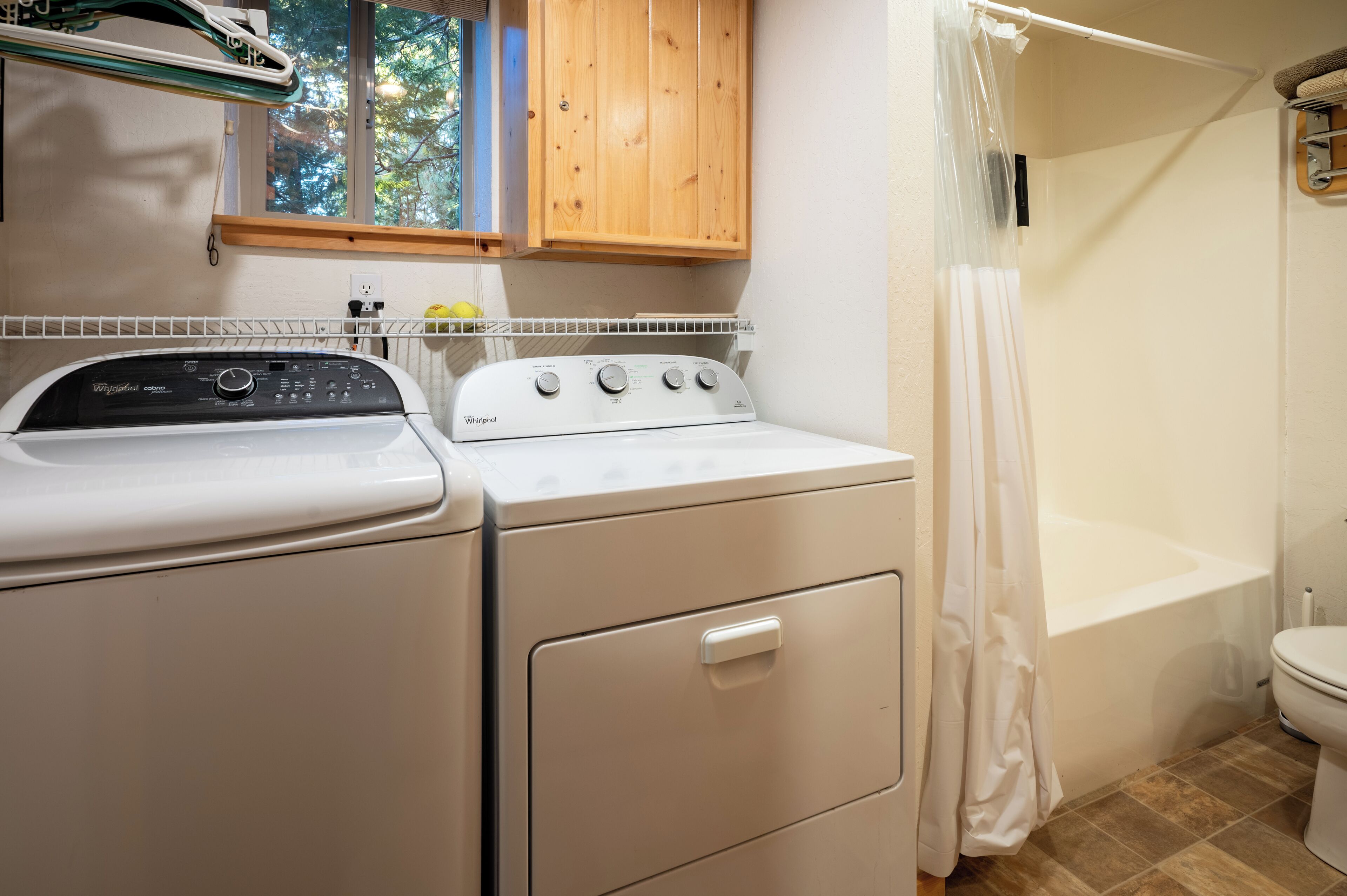 Un espace buanderie comprenant un lave-linge et un sèche-linge à chargement frontal, un meuble en bois au-dessus, et une douche avec un rideau.
