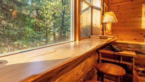 A wooden cabin interior with a large window overlooking a forest, a wooden table with a lamp, and a telephone.