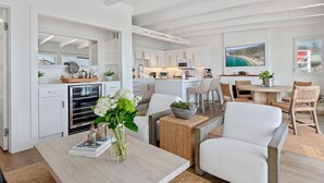 Une cuisine moderne avec des meubles blancs, un coin repas avec une table ronde et des chaises en osier, et un salon avec un canapé blanc et une table basse.