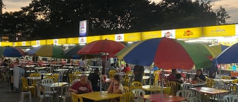 An outdoor dining area with yellow and white plastic chairs and tables, some under colorful umbrellas.
