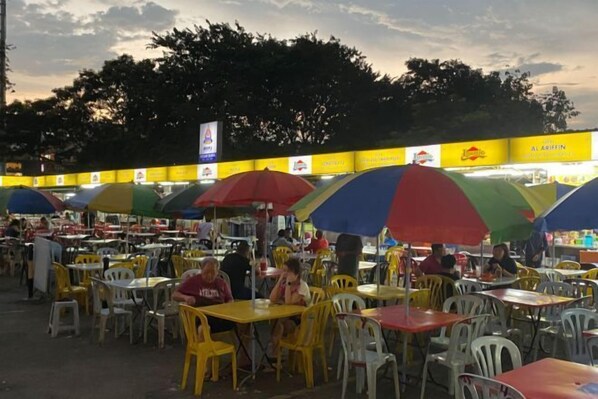 Un espace extérieur aménagé pour les repas, avec des chaises et des tables en plastique jaune et blanc, dont certaines sont placées sous des parasols colorés.