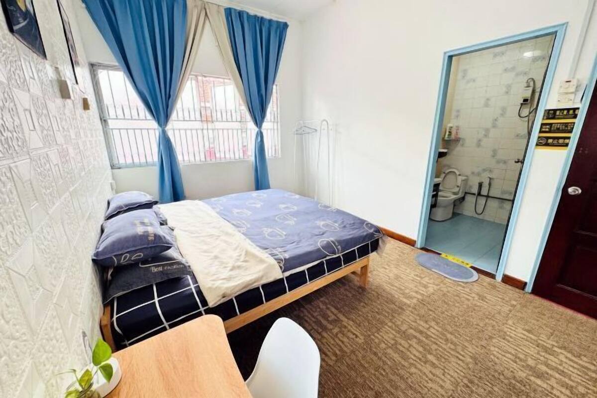 A bedroom with a bed, a desk, a chair, a plant, and a bathroom visible through an open door.