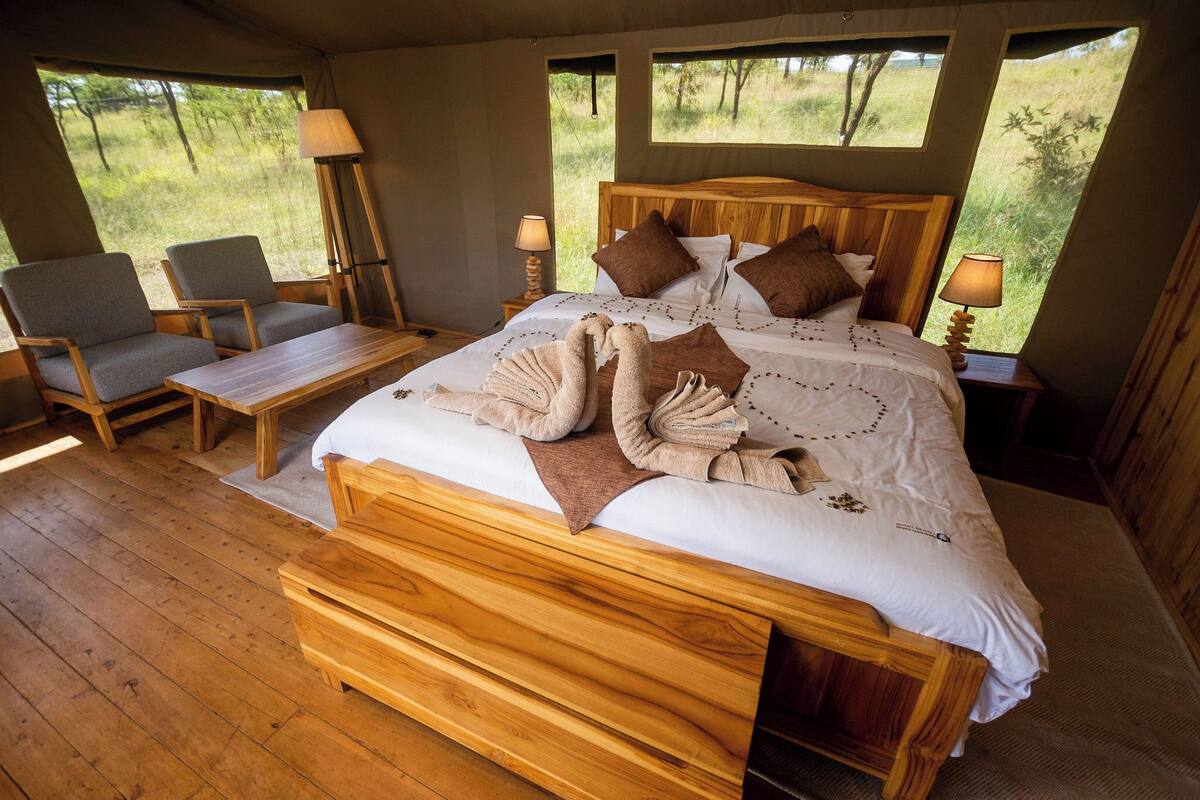A spacious bedroom with a large bed, wooden headboard, two bedside tables with lamps, a wooden bench, and two chairs.