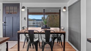 A modern dining area with a wooden table and black chairs, a potted plant, and a view of a lake and mountains through large windows. - Lakefront Home with Private Beach and Dock | Outdoor Dining (Harrison)