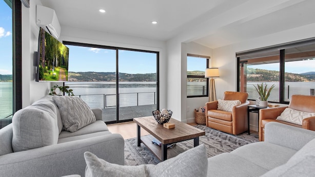 A modern living room with a large window overlooking a lake, a flat-screen TV mounted on the wall, and a cozy seating area with leather sofas and a wooden coffee table. - Lakefront Home with Private Beach and Dock | Outdoor Dining (Harrison)