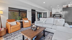 A modern living room with a sofa, two leather chairs, a wooden coffee table, and a view of the kitchen. - Lakefront Home with Private Beach and Dock | Outdoor Dining (Harrison)