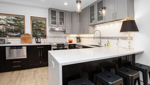 A modern kitchen with dark cabinets, a white island, bar stools, stainless steel appliances, and pendant lighting.